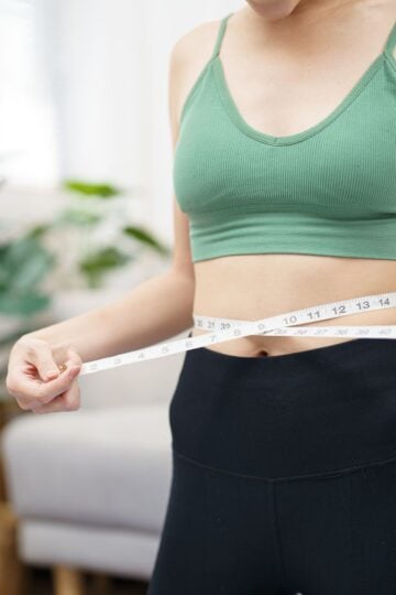 A person in a green top and black pants measures their waist with a measuring tape, perhaps assessing progress on their weight loss journey. A plant and couch are in the background, adding a touch of calm to the scene.