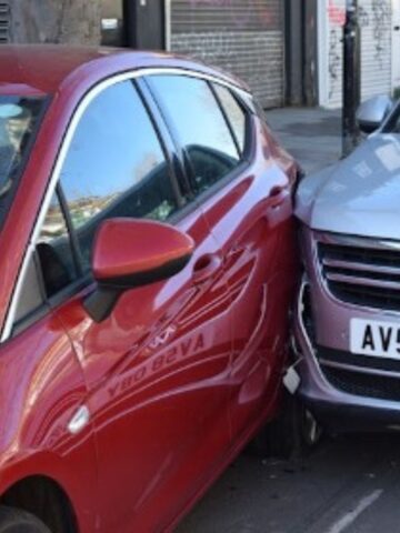 After a car accident, a red car and a silver car collide on the street, with the front of the silver car pressing against the side of the red one.