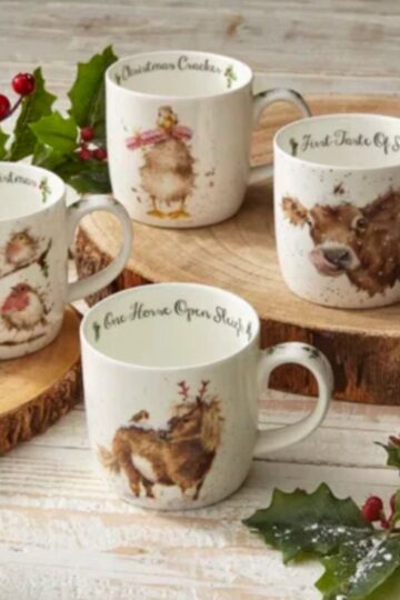 Four festive mugs with holiday designs and charming phrases, including animals and holly, make the perfect Christmas gift. They are beautifully displayed on a wooden surface surrounded by rustic holiday decorations, highlighting their appeal as quality homeware.