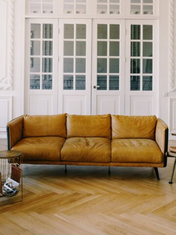 How To Clean A Faux Leather Couch (Care Guide) A tan faux leather sofa sits in a bright room with parquet flooring, accompanied by a wire side table and a wooden chair. White paneled doors with glass sections grace the background, while easy-care maintenance ensures the couch stays pristine.