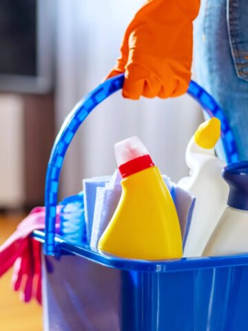 A person equipped for a deep clean holds a blue bucket brimming with cleaning supplies, including spray bottles and cloths, all while donning orange gloves. It's a perfect winter task to freshen up your space and tackle those overlooked things to clean.