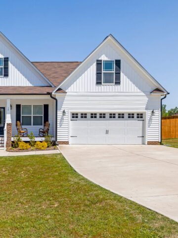 Single-story white house with brown roof, attached two-car garage, and concrete driveway that could benefit from professional driveway cleaning. Two small trees are planted in the front yard next to the porch, with a fence visible in the background. Perfect for those considering hiring maintenance services.