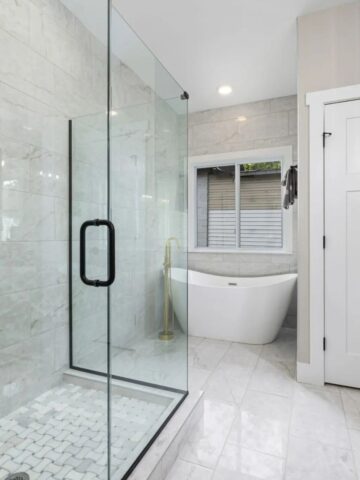 A modern bathroom featuring a sleek glass-enclosed walk-in shower and a freestanding bathtub. The white tiled walls and floor elevate the design, complemented by a window next to the tub.