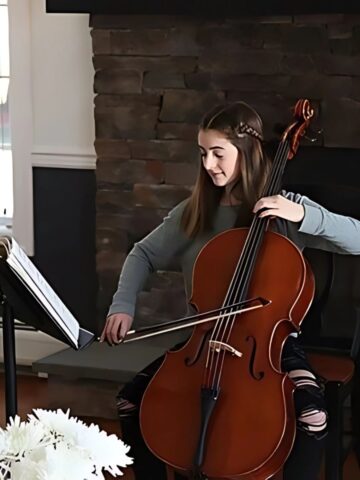 A well-rounded individual, the woman plays the cello next to a music stand in a cozy room with a stone fireplace, highlighting her passion for music.