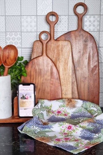 A selection of wooden cutting boards and a utensil holder with wooden utensils, alongside a smartphone on a stand and a floral napkin, are stylishly arranged on a kitchen counter with a tiled backsplash.