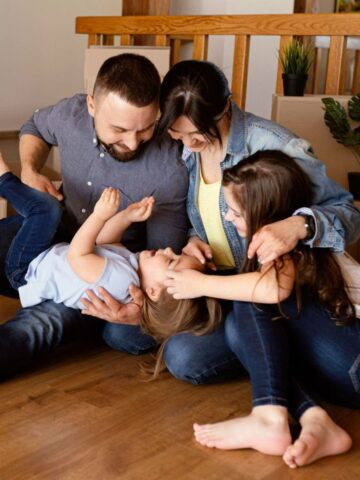 A family of four sits on the floor among boxes, enjoying the warmth and joy of their new home. The parents play with their young children, creating an atmosphere of family comfort in a space that promises a lifetime of home safety.