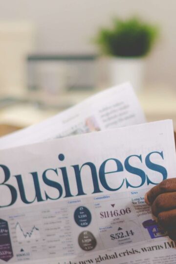 A person is intently reading a newspaper section titled "Business," with stock prices and charts in clear view. A blurred potted plant graces the background, offering a serene touch as they explore proven strategies for building a thriving business.