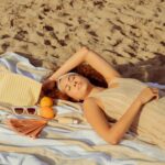 A woman in stylish swimwear lounges on a beach towel, eyes closed. Beside her are summer must-haves: chic sunglasses, juicy oranges, a fan for the heat, and a striped beach bag holding a refreshing bottle.