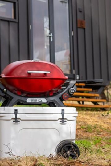 A red portable charcoal grill sits atop a white cooler outdoors, with a small dark-colored cabin in the background.