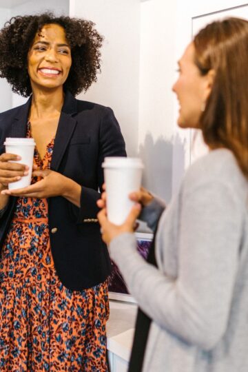 Two people stand indoors with coffee cups, engaged in a networking conversation. One person has curly hair and wears a blazer over a floral dress, while the other, possibly a freelancer, has long straight hair and a cardigan.