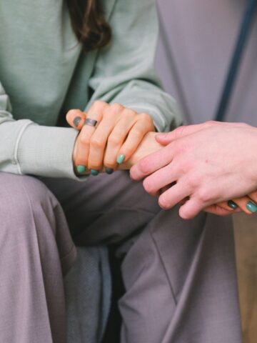 Two people sit close together holding hands, one wearing a green sweater and gray pants, both with green nail polish.