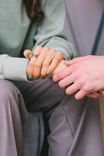 Two people sit close together holding hands, one wearing a green sweater and gray pants, both with green nail polish.