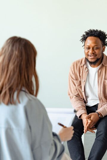 In a softly lit room with light walls, a man sits on a chair facing a woman who holds a pen and clipboard, discussing an affordable psychiatry service in Los Angeles.