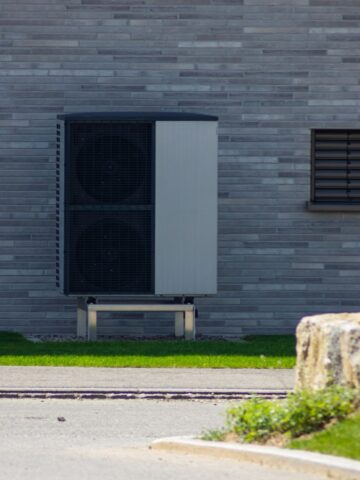 How to Protect Your Heat Pump from Weather and Debris An outdoor air conditioning unit, featuring a durable weather protection cover, is mounted on a metal stand against a gray brick wall with a small window on the right.