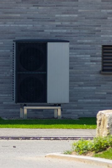 An outdoor air conditioning unit, featuring a durable weather protection cover, is mounted on a metal stand against a gray brick wall with a small window on the right.