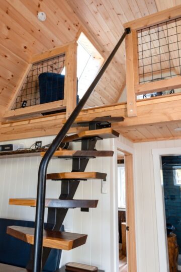 Wooden loft interior with spiral staircase showcases a stunning loft transformation. Upper area secured with wire railing, while the lower area offers seating with a black and white cushion. Functional living space is bathed in sunlight filtering through skylights.