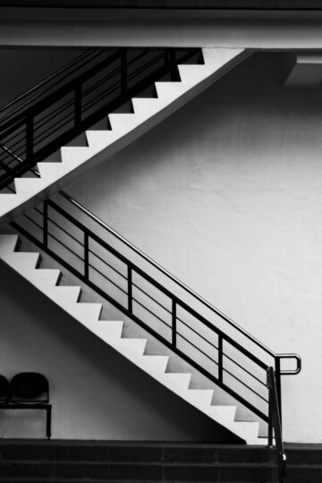 Black and white image of a staircase hinting at structural issues, with a metal railing against a plain wall. Two empty chairs sit at the bottom on a landing, their placement suggesting silent conversations about hidden cracks or unseen staircase problems.