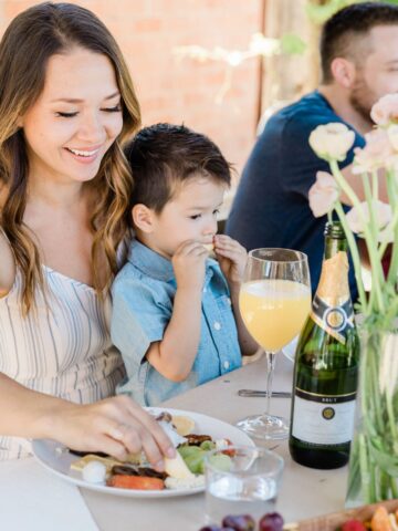 A woman and child enjoy a delightful brunch at an outdoor table, adorned with flowers and a bottle. A man and another child play in the background, capturing the essence of a serene Mother's Day celebration at home.