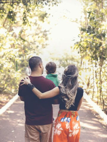 A family of three, embodying important parenting tips, strolls down a tree-lined path. With their child nestled between the adults, they walk confidently surrounded by lush green foliage, showcasing the essence of smart parenting in each step they take.