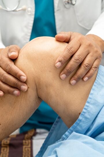 A healthcare professional examines a patient's knee, gently pressing and supporting the joint while the patient lies on an examination table covered with a blue drape; assessment may precede procedures like a sacroiliac joint injection.