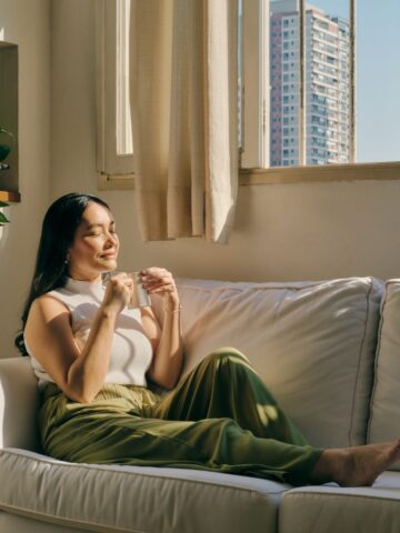 Mindfulness on the Move: Pocket-Sized Tools to Manage Anxiety Anywhere Woman sitting on a white couch by a window, holding a cup and enjoying sunlight, practicing mindfulness as buildings are visible outside.