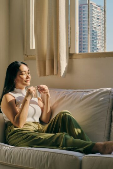 Woman sitting on a white couch by a window, holding a cup and enjoying sunlight, practicing mindfulness as buildings are visible outside.