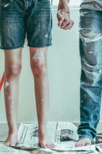 Two people are standing barefoot on newspaper, holding hands. Both wear paint-stained clothes and jeans—one holds a paintbrush, ready for some home maintenance. A broom on the floor nearby hints at a low-stress cleanup for busy families.
