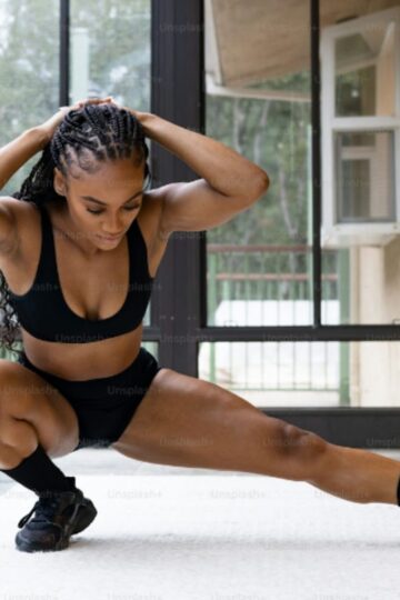 A woman in athletic wear performs a side lunge stretch indoors on a light-colored floor near large windows, showcasing the flexibility that training with a Sculptformer versatile machine can provide.