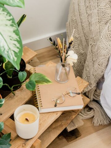 A wooden bedside table with small comforts: a potted plant, beige notebook, eyeglasses, lit candle, and a small vase of dried flowers beside a knitted blanket-perfect to help you recharge after a long day.