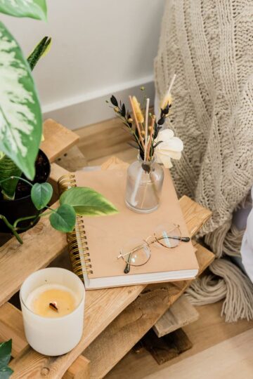 A wooden bedside table with small comforts: a potted plant, beige notebook, eyeglasses, lit candle, and a small vase of dried flowers beside a knitted blanket—perfect to help you recharge after a long day.