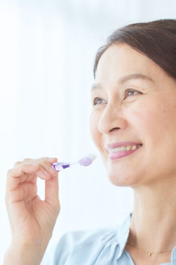 A woman with sensitive teeth smiles while brushing her teeth with a purple toothbrush.