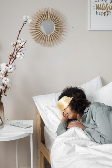 After a long workday, a person wearing a gold sleep mask is lying in bed under white blankets beside a nightstand with a vase, notebook, and speaker in a minimalist bedroom to de-stress naturally.