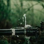 Water sprays from a leak in a black metal pipe outdoors, with green vegetation in the blurred background-a clear sign of a plumbing emergency requiring immediate attention.