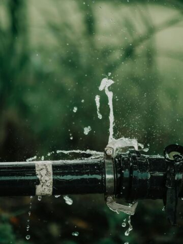 Water sprays from a leak in a black metal pipe outdoors, with green vegetation in the blurred background-a clear sign of a plumbing emergency requiring immediate attention.