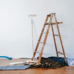 A wooden ladder, paint roller, and painting supplies are set up against a white wall, with drop cloths and painter's tape protecting the floor during a home improvement project.