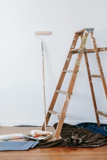 A wooden ladder, paint roller, and painting supplies are set up against a white wall, with drop cloths and painter’s tape protecting the floor during a home improvement project.