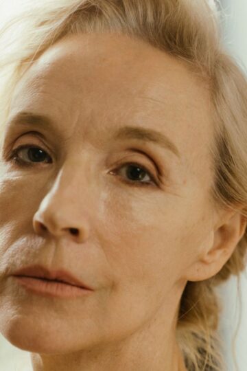Close-up of an older woman with light skin and blond hair, looking directly at the camera with a neutral expression. Her face wrinkles are visible, captured naturally against a softly blurred background.