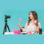 A self-employed blogger sits at a table with makeup items, holding social media like icons, facing a camera on a tripod with a blue background, ready to share her latest blogging tips.