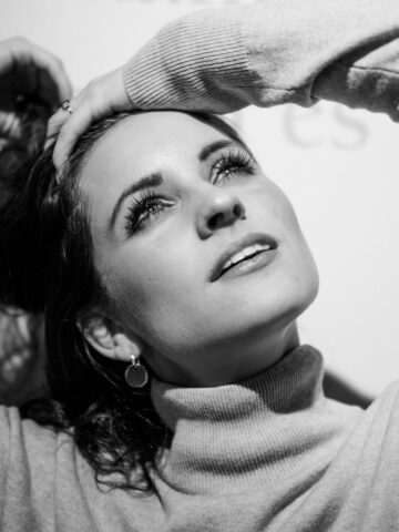 Black and white photo of a woman in a turtleneck gazing upward, showcasing a modest look with one hand on her head and the other behind her hair.