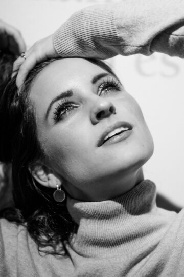 Black and white photo of a woman in a turtleneck gazing upward, showcasing a modest look with one hand on her head and the other behind her hair.