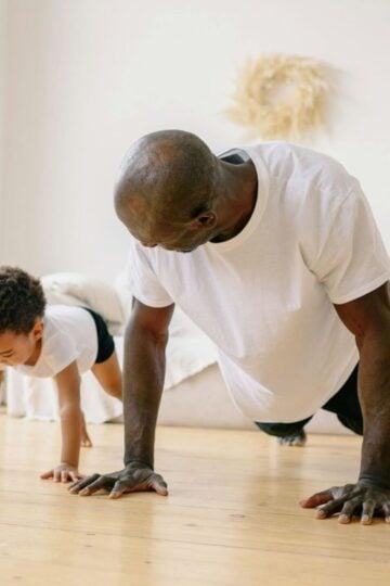 A busy dad and his child do push-ups together on a wooden floor in a bright living room, celebrating fitness and fatherhood.