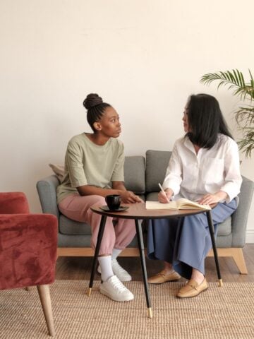 Admitting You're Addicted: Five People To Talk To Two women sit on a sofa having a conversation about admitting addiction; one listens while the other writes in a notebook on the coffee table between them. A red chair and a potted plant are nearby.