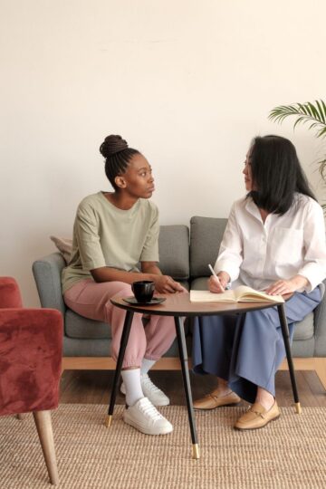 Two women sit on a sofa having a conversation about admitting addiction; one listens while the other writes in a notebook on the coffee table between them. A red chair and a potted plant are nearby.
