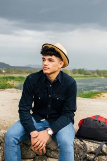 A person in a black shirt and jeans sits on a stone wall by a river, exuding travel energy with their straw hat. Beside them rests a red and black backpack, ready for the journey ahead under the cloudy sky.