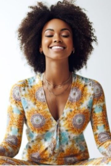 Person with curly hair smiling and sitting cross-legged in a patterned outfit, meditating against a plain background.