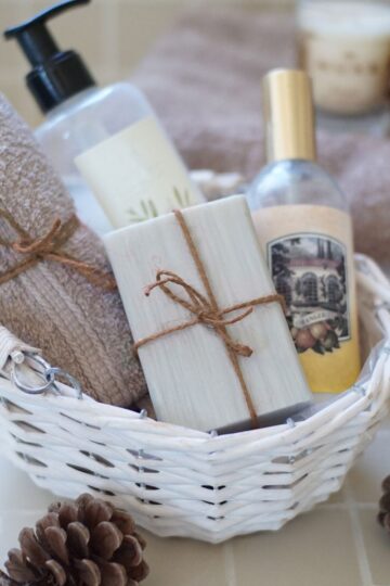 White wicker basket with beauty staples like a towel, soap, lotion, and a spray bottle tied with twine, placed on a tiled surface. Brown pine cones and a folded towel in the background complete the serene everyday beauty routine setting.