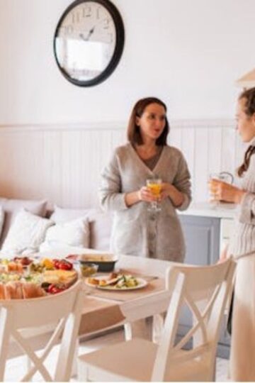 Two women stand by a dining table with a large spread of food, talking and holding drinks. A clock is mounted on the wall behind them. The room's cozy, light-filled ambiance sets the perfect scene for relaxing home parties.