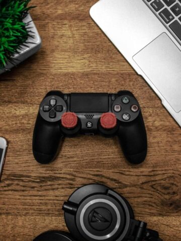 A game controller rests on a wooden desk, surrounded by a laptop, smartphone, headphones, and a small plant-perfect companions for a lazy day indulging in classic games.