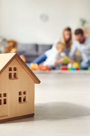 A wooden toy house is in focus in the foreground, embodying a safety-first approach, while a family joyfully plays together out of focus in the background.