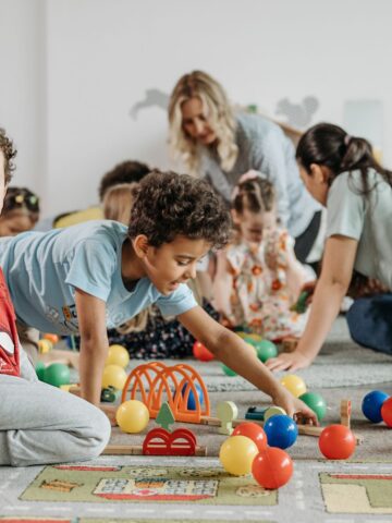 Children play with colorful balls and toys on a carpet, while adults supervise in the background-a scene right out of a parent's guide to choosing childcare wisely.